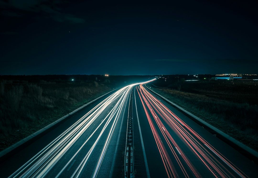 Eine nächtliche Autobahn mit mehreren Spuren, beleuchtet von Scheinwerfern und Rücklichtern, die sich unter einem dunklen Himmel in die Ferne erstreckt.
