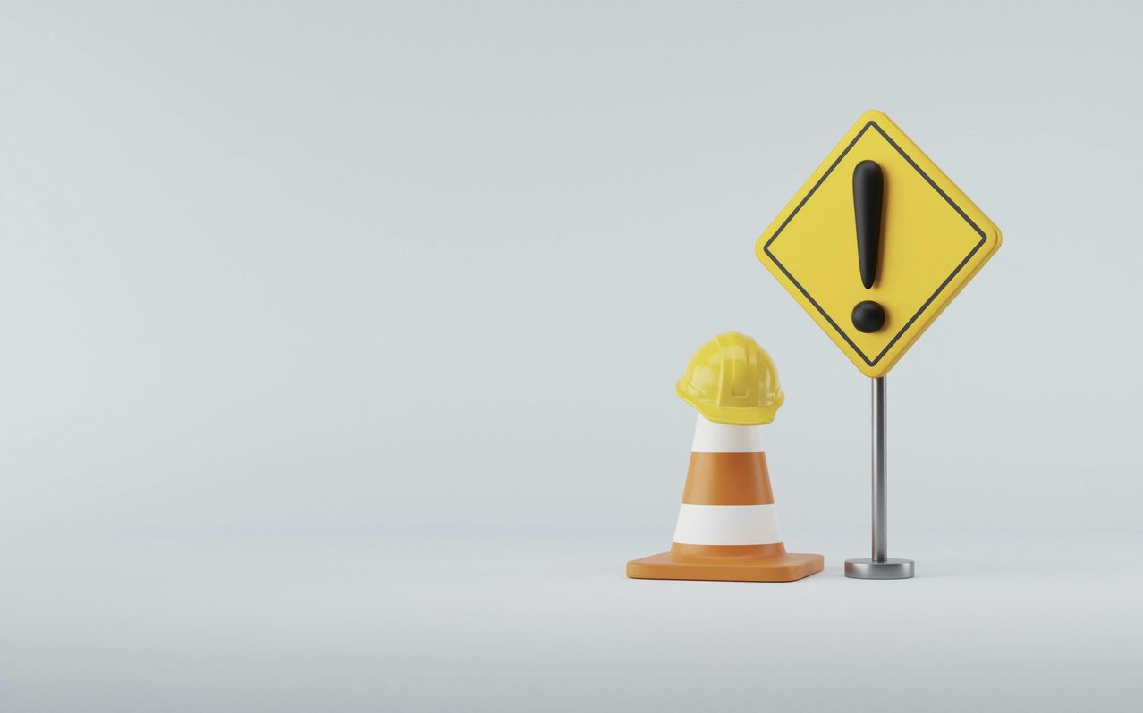 Ein gelbes Warnschild mit schwarzem Ausrufezeichen steht neben einem Verkehrskegel mit einem gelben Schutzhelm obenauf.