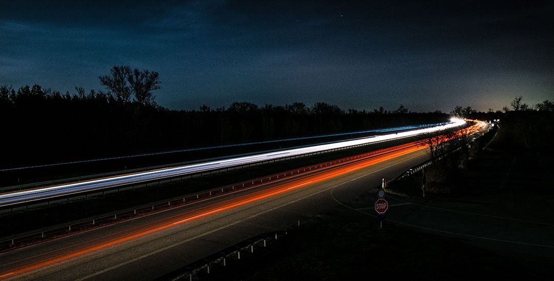 Eine Nachtautobahn mit langen Streifen von Scheinwerfern und Rücklichtern, die auf schnelle Fahrzeuge hindeuten. Ein Stoppschild ist an der Seite sichtbar.