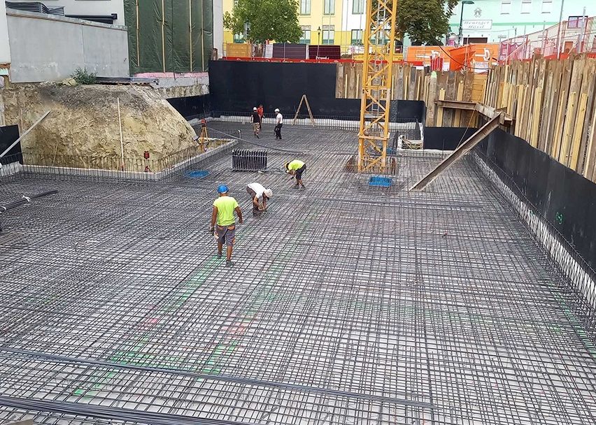 Bauarbeiter arbeiten an einem Betonfundament mit einem Stahlbewehrungsgitter. Ein Haufen Erde ist in der Nähe, und ein gelber Kran ist im Hintergrund.