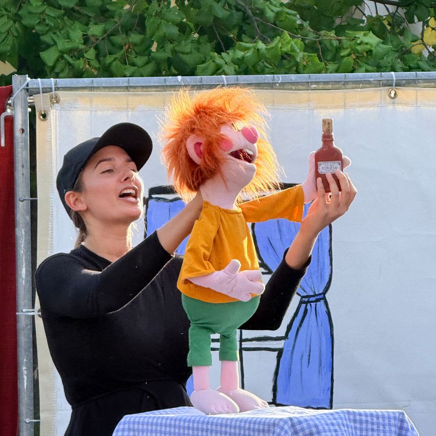 Eine Frau hält eine Puppe mit orangefarbenen Haaren und einem gelben Shirt, sie lacht und hält eine Flasche Medizin.