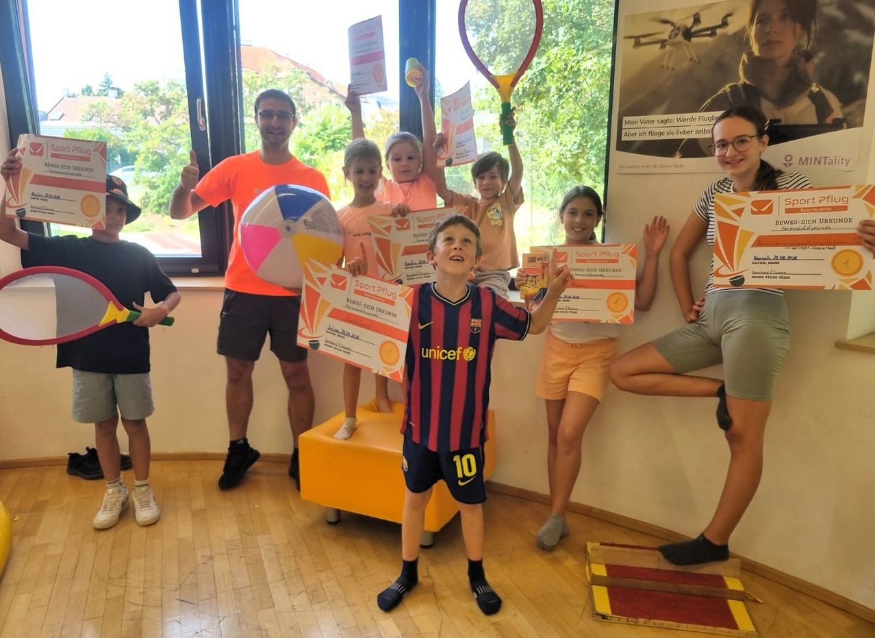 Eine Gruppe von Kindern und Erwachsenen steht in einem Raum, hält Urkunden und lächelt. Der Junge in der Mitte trägt ein Barcelona-Fußballtrikot. Hinter ihnen ist ein Glasfenster mit Blick auf Bäume.