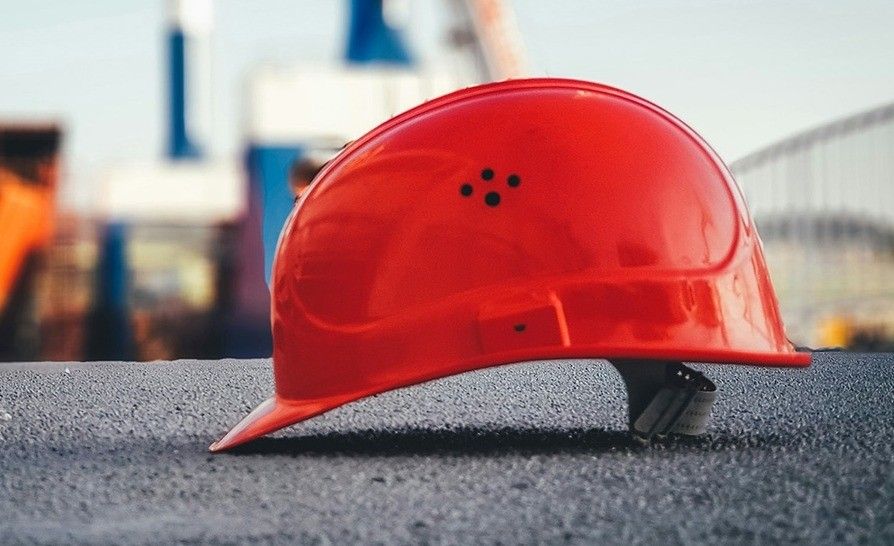 A closeup of a red hard hat on a grey surface. Behind it is a blurry background with a person standing near a blue structure.