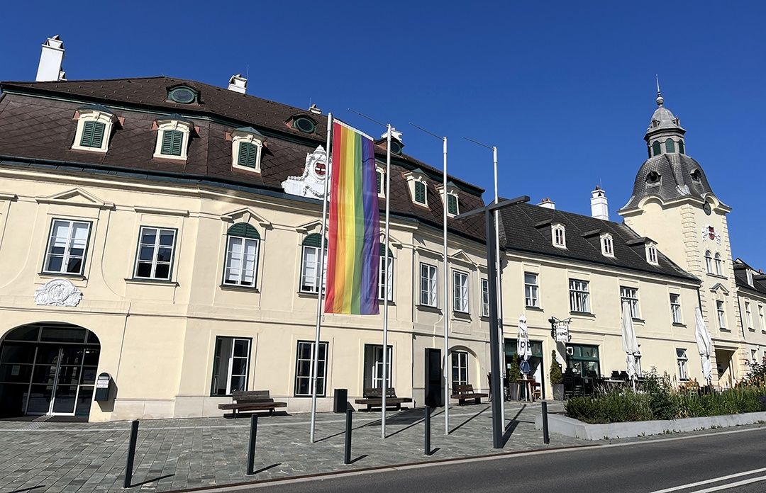 A large beige building with many windows and a brown roof. A rainbow flag flies in front. There are benches and potted plants outside.