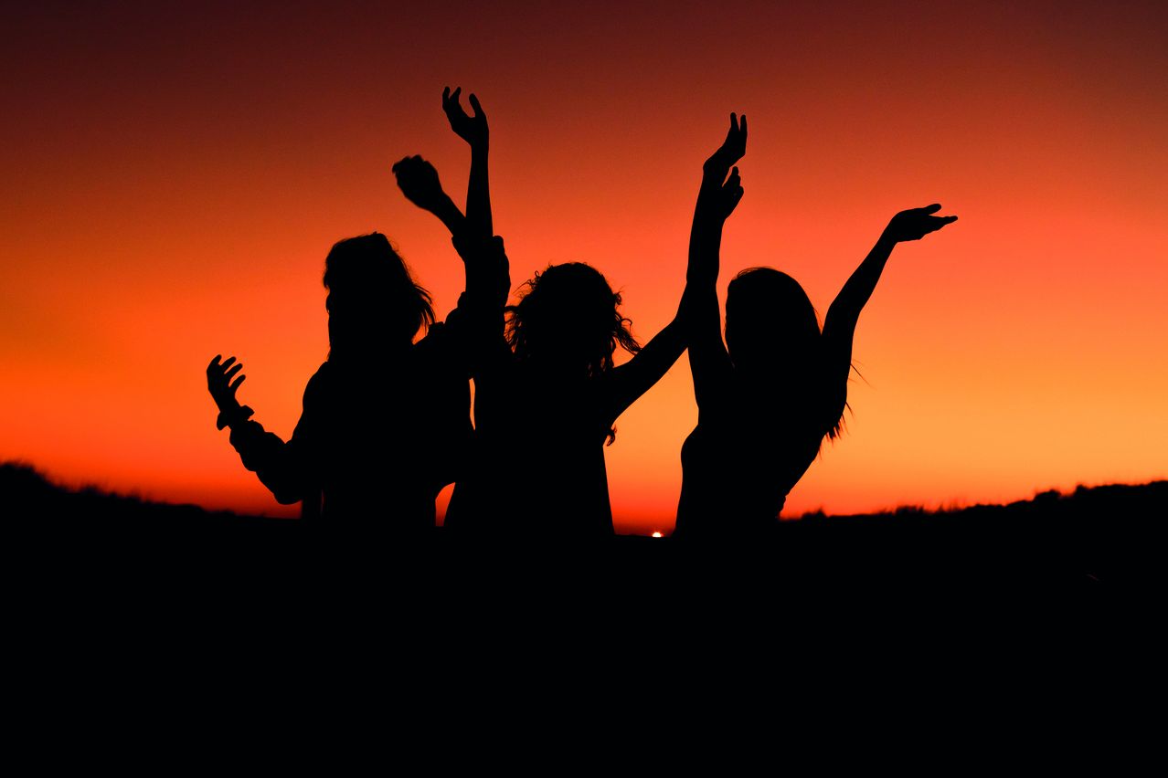 Three silhouetted figures raise their arms in the sunset. They stand against a backdrop of vibrant orange and red hues.
