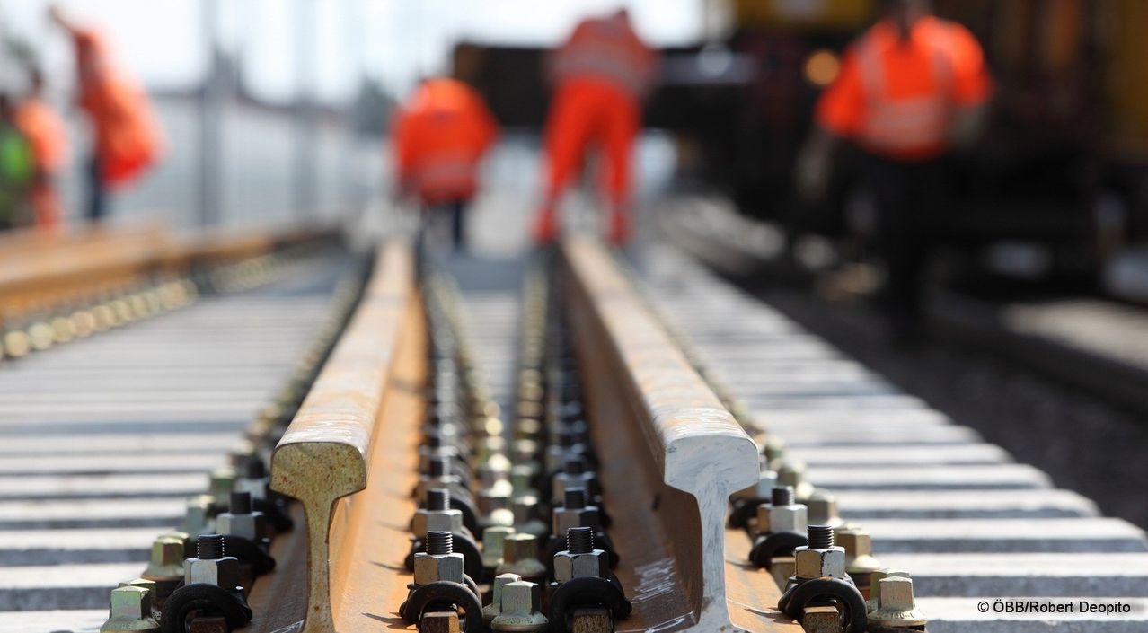 Eine Nahaufnahme einer Eisenbahnschiene mit Bolzen und Metallstücken im Fokus, während Arbeiter in orangefarbenen Westen im Hintergrund verschwommen sind.