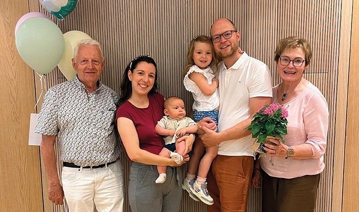 Eine Familie mit fünf Personen steht lächelnd vor einer Holzwand. Der Mann links trägt einen Gürtel, während die Frau rechts einen Blumenstrauß hält. Hinter ihnen sind drei Ballons an der Wand befestigt.
