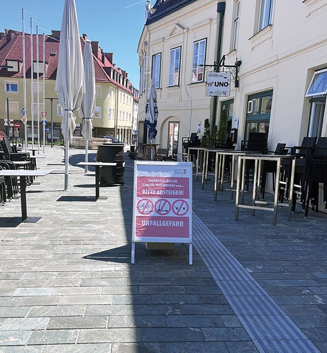 Eine Außengastro mit leeren Tischen und Stühlen unter Schirmen. Ein Schild weist darauf hin, dass Roller und Skateboards verboten sind. Das Café heißt 'UNO'.