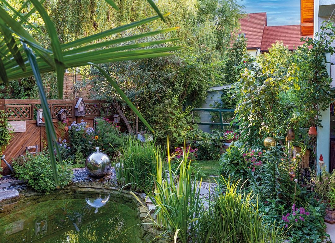 A garden with a pond in the center, surrounded by flowers, plants, and a silver globe. Trees and a wooden fence are visible in the background.