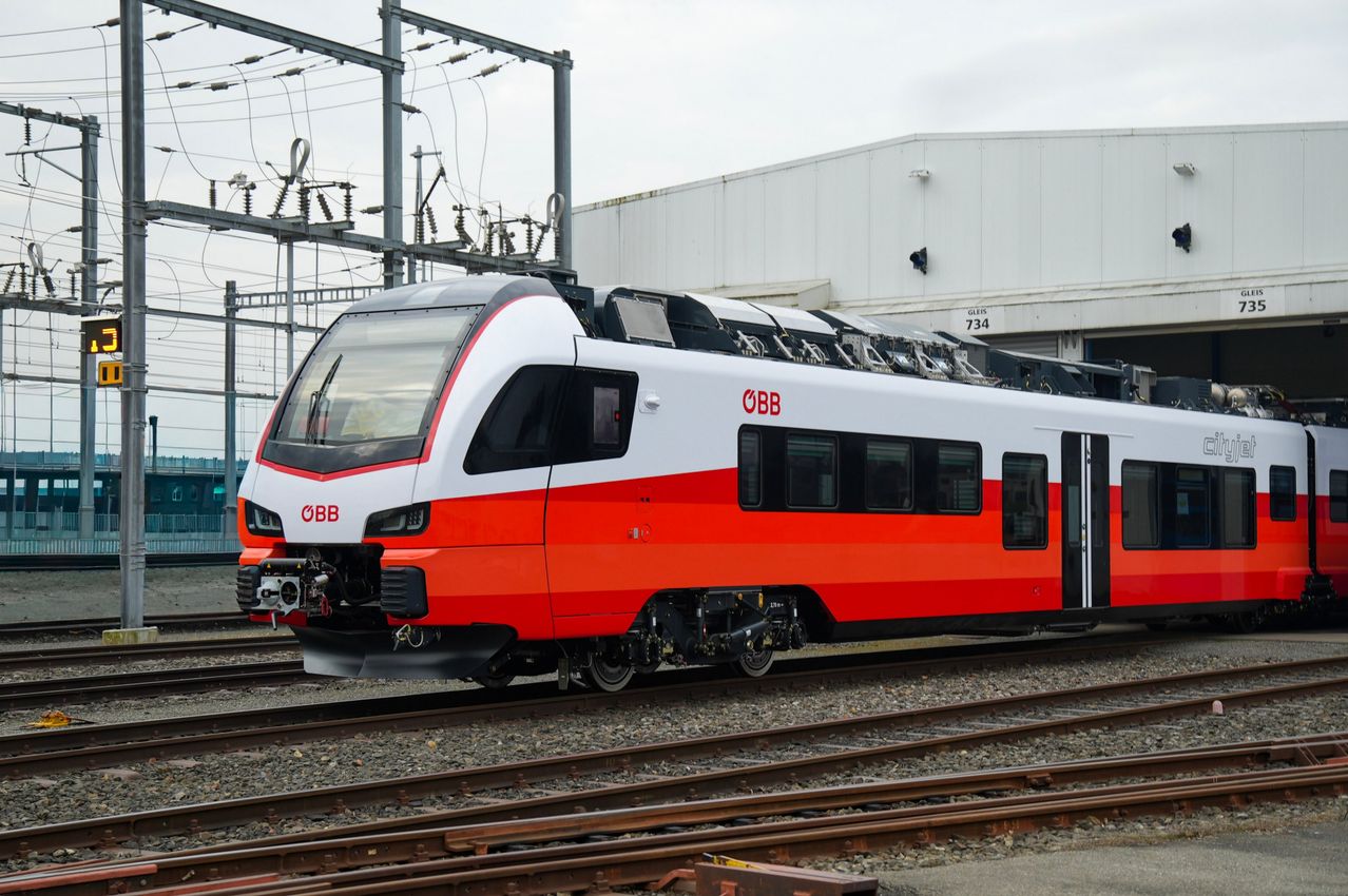 Ein orange-weißer Zug mit den Buchstaben 0BB steht auf den Bahngleisen. Er steht vor einem Gebäude mit der Nummer 734.