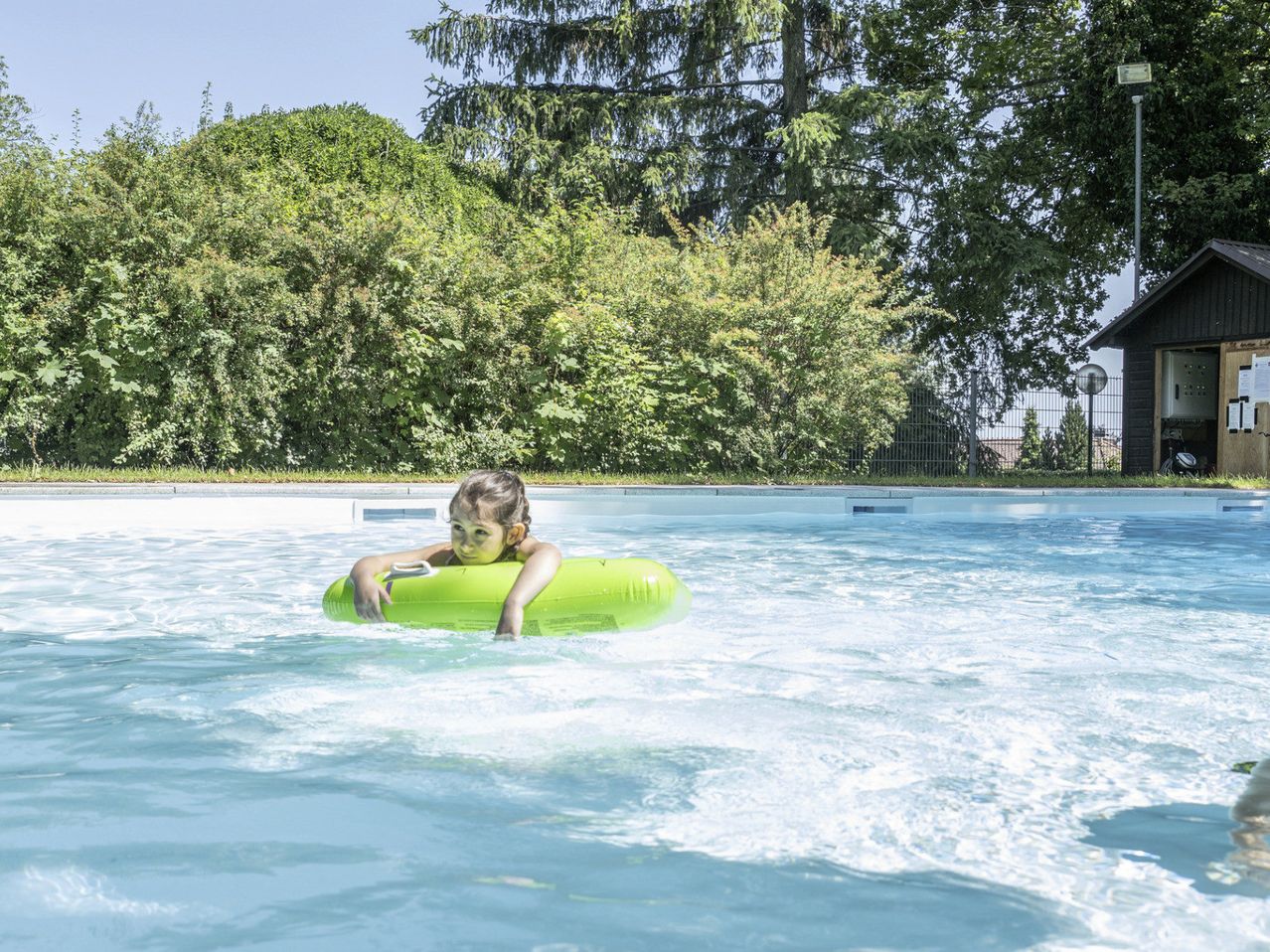 Ein junges Mädchen schwimmt in einem Pool mit einem grünen Schwimmreifen unter einem strahlend blauen Himmel, mit Bäumen im Hintergrund.