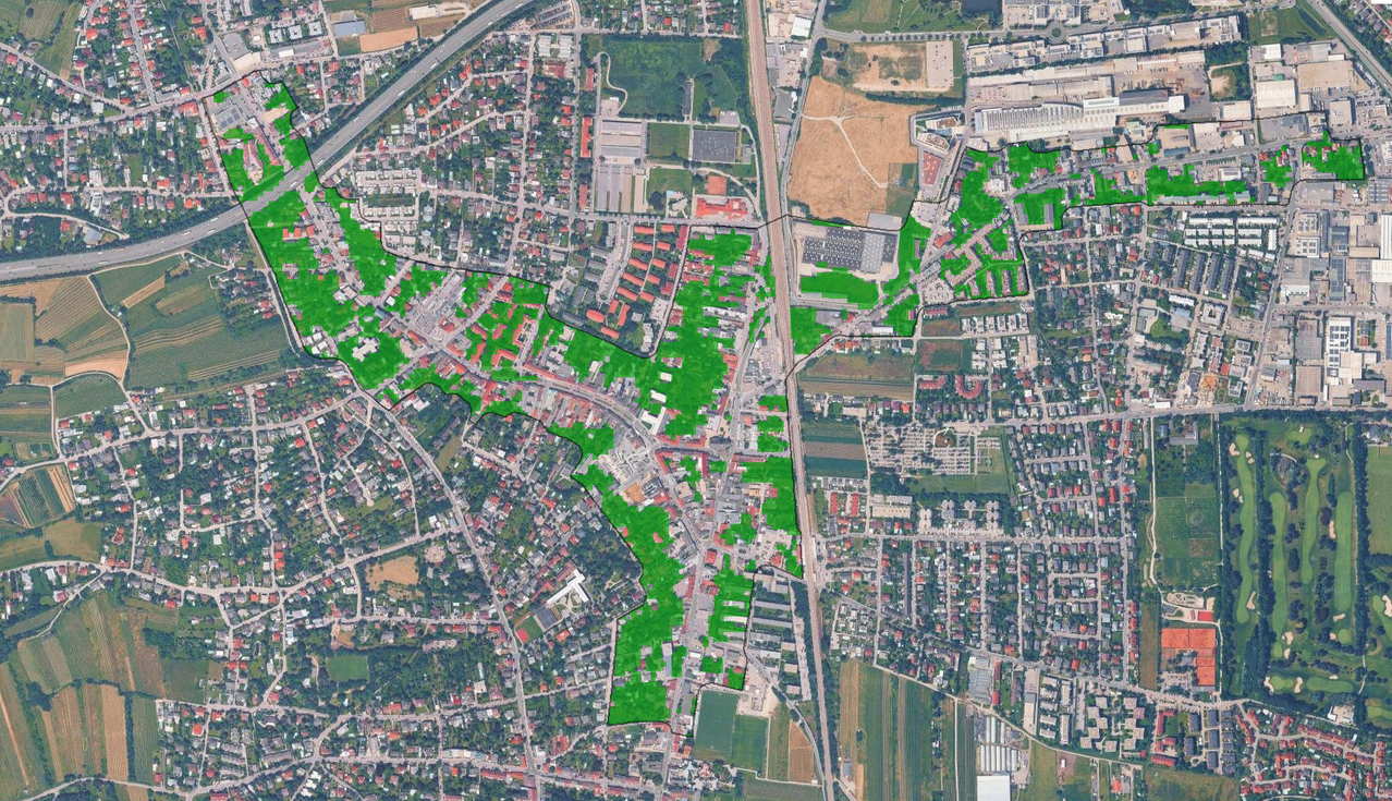 Luftbild einer Stadt mit grünen Überlagerungen, die bestimmte Bereiche markieren, umgeben von Straßen und offenen Feldern. Gebäude sind in verschiedenen Größen und Stilen, einige mit roten Dächern.