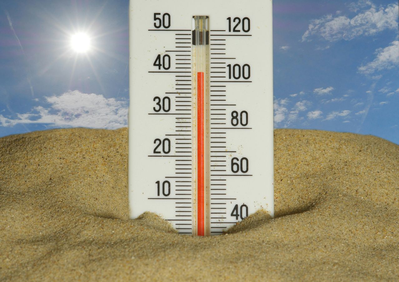 Ein Thermometer, das im Sand vergraben ist und eine Temperatur von 100 Grad Fahrenheit anzeigt, mit einem sonnigen Himmel im Hintergrund.