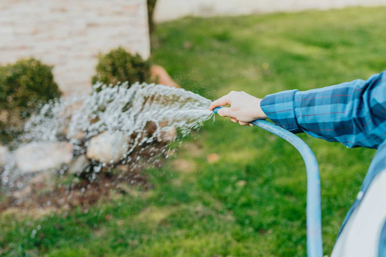 Eine Person bewässert einen Garten mit einem blauen Schlauch, wobei Wasser auf einen grünen Rasen spritzt.