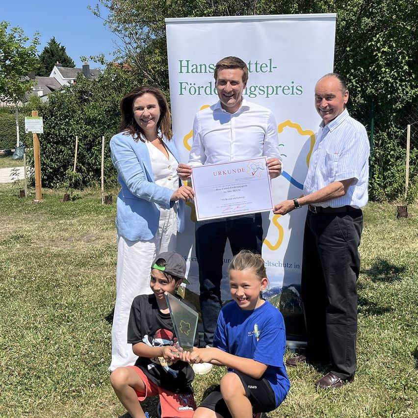 Eine vierköpfige Familie, darunter zwei Kinder, posiert mit einem Auszeichnungszertifikat und einem Pokal auf einem Rasen, vor einem Banner im Hintergrund.