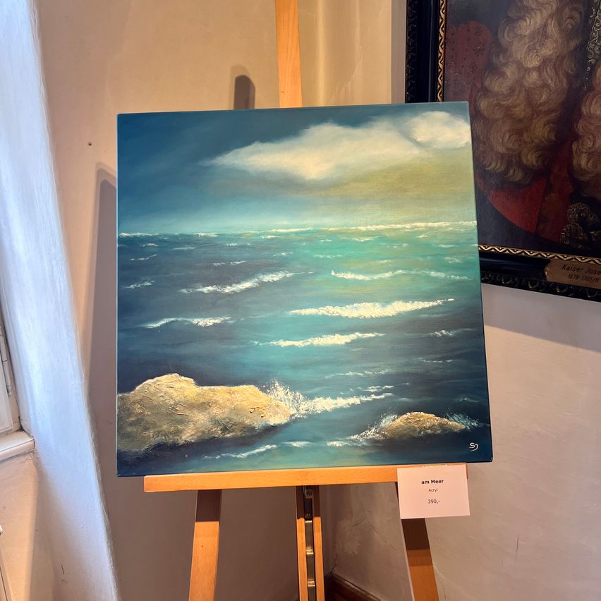 Ein Kunstgemälde einer Ozeanszene mit Wellen, die auf Felsen brechen. Es steht auf einem Staffelei mit einem Schild, das 'am Meer' und einen Preis von 390.- anzeigt.