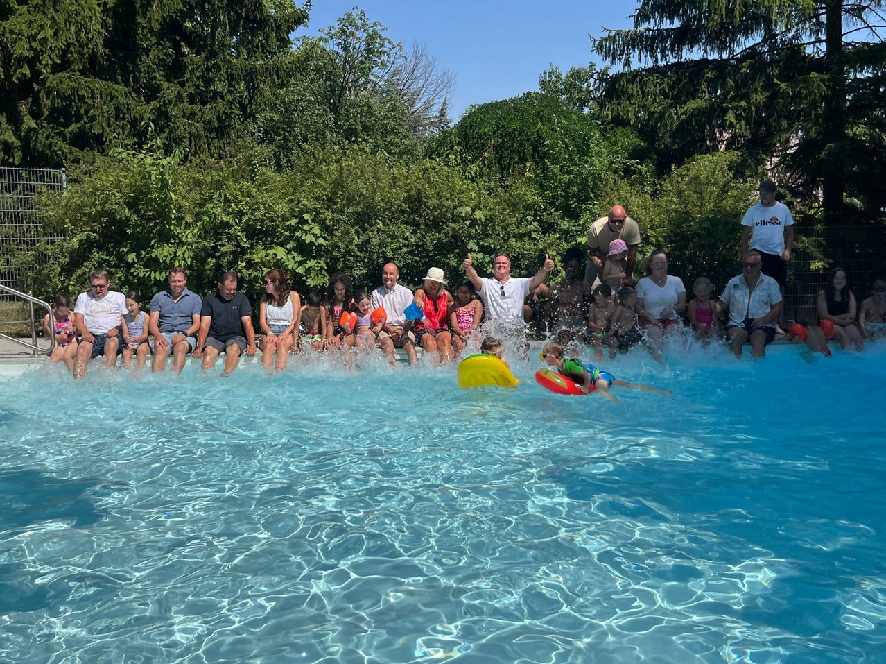 Eine Gruppe von Erwachsenen und Kindern sitzt am Rand eines Pools und feuert zwei Kinder im Wasser an. Die Kinder tragen Schwimmhilfen und eines taucht ins Wasser ein.