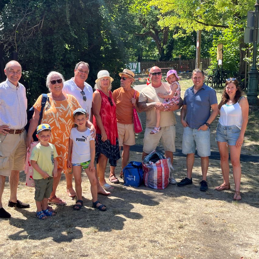 Eine Familie von acht Personen, bestehend aus Erwachsenen und Kindern, posiert für ein Foto in einem Park mit Bäumen und einem Holzzaun im Hintergrund.