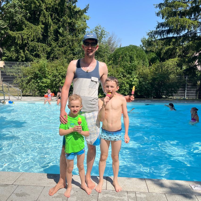 Ein Mann und zwei Jungen stehen in einem Schwimmbad an einem sonnigen Tag. Sie halten Eis am Stiel in den Händen.