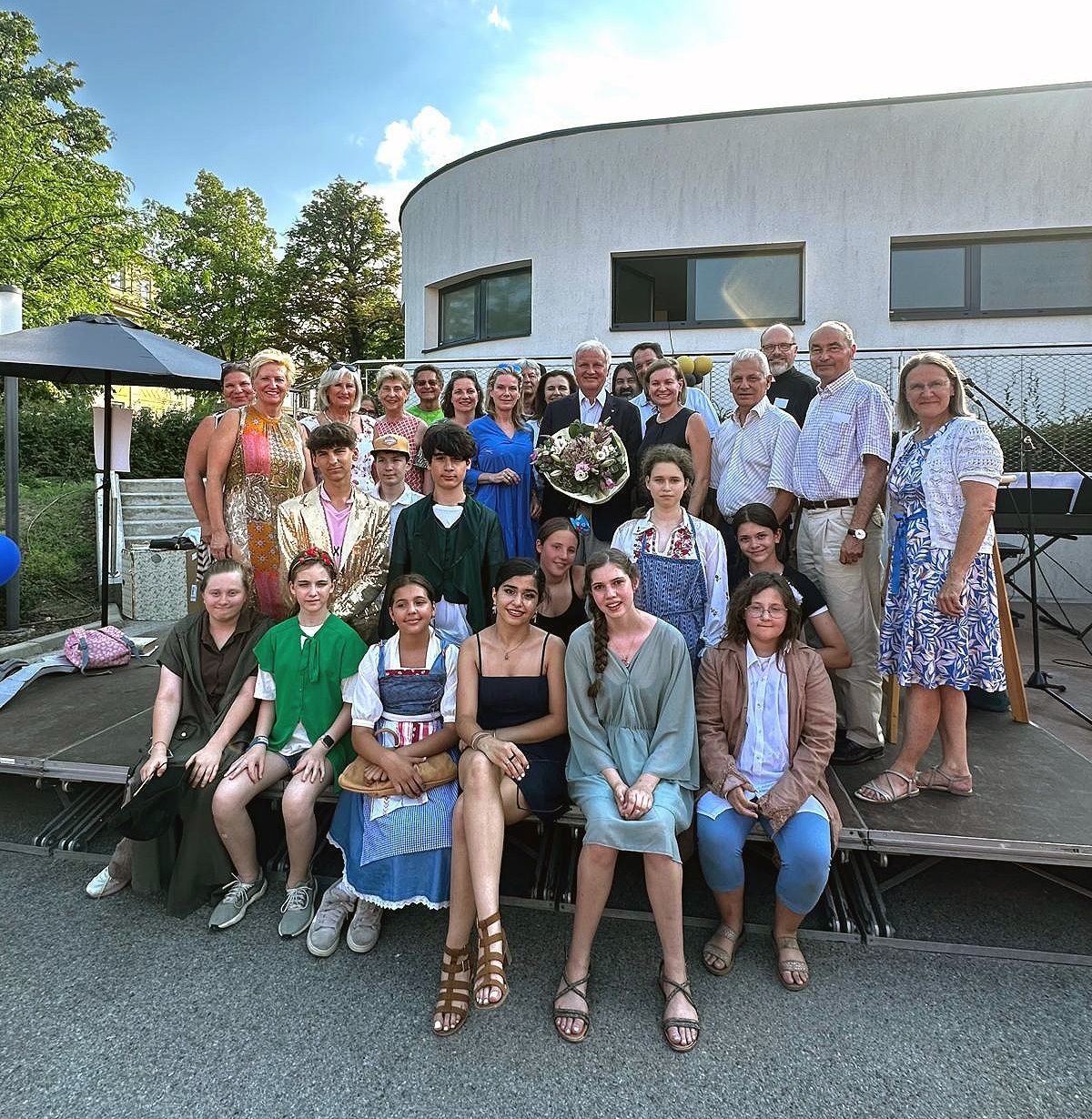Group photo of a diverse gathering of people, some seated and some standing, in front of a modern building. Trees and a large umbrella are visible in the background.