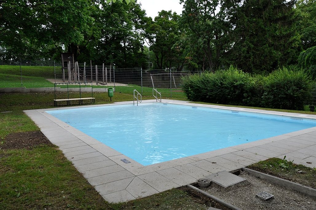 An empty swimming pool surrounded by a concrete walkway, with benches and a green trash bin nearby, set in a park with lush trees and bushes.