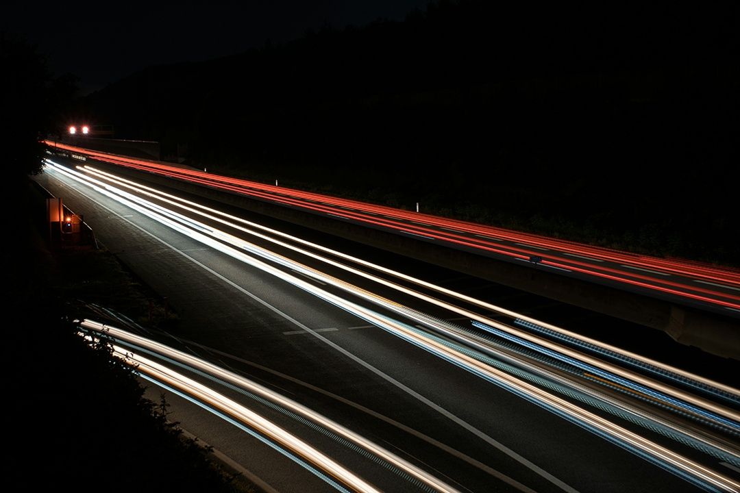 Eine Nachtautobahn mit mehreren Spuren, beleuchtet von Autoscheinwerfern, die lange Lichtspuren erzeugen. Die Straße ist leer, und die Dunkelheit wird nur von den Lichtstreifen durchbrochen.