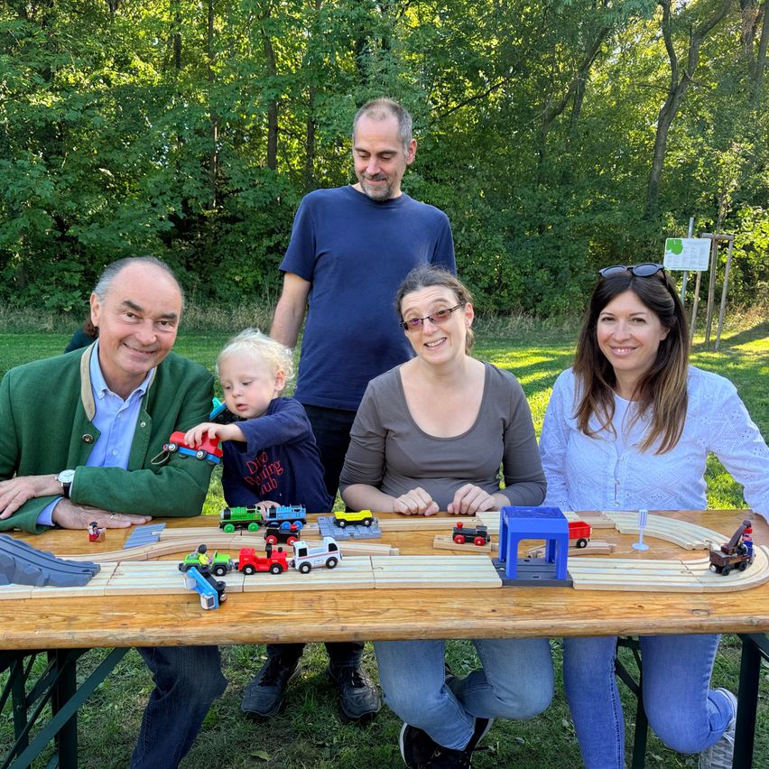 Familie spielt mit Holzeisenbahn im Garten, Mann im grünen Sakko, Kind, Frau, Mann, Frau