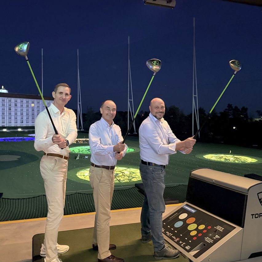 Drei Männer stehen in einem Golfsimulator und halten Golfschläger. Sie lächeln und posieren für ein Foto.
