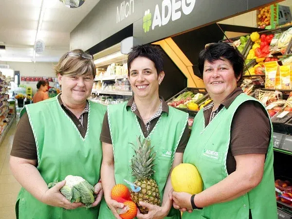Drei Frauen in grünen Westen stehen in einem Lebensmittelgeschäft und halten Obst und Gemüse. Hinter ihnen ist ein Regal mit verschiedenen Obst- und Gemüsesorten zu sehen, und ein Schild mit der Aufschrift 'ADEG' ist an der Wand angebracht.