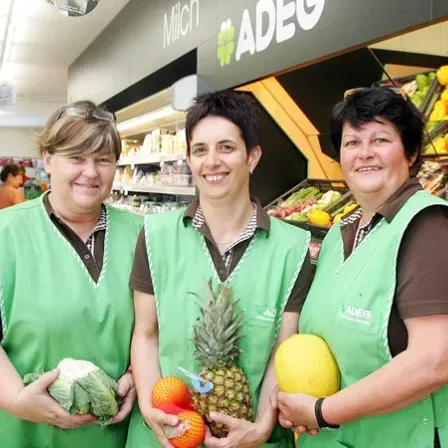 Drei Frauen in grünen Westen stehen in einem Lebensmittelgeschäft und halten Obst und Gemüse. Hinter ihnen ist ein Regal mit verschiedenen Obst- und Gemüsesorten zu sehen, und ein Schild mit der Aufschrift 'ADEG' ist an der Wand angebracht.