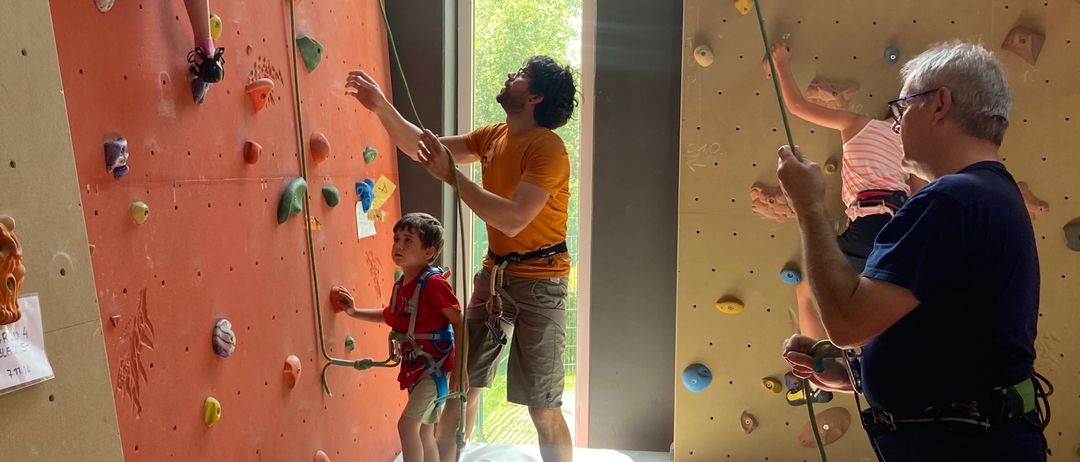 Mehrere Personen sind in einem Fitnessstudio mit dem Klettern beschäftigt. Ein Mann in einem gelben T-Shirt hilft einem kleinen Jungen an der Kletterwand. Ein älterer Mann in einem blauen T-Shirt beobachtet. Ein Mädchen klettert ebenfalls an der Wand, hinter ihr steht eine weitere Person.