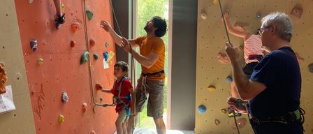 Mehrere Personen sind in einem Fitnessstudio mit dem Klettern beschäftigt. Ein Mann in einem gelben T-Shirt hilft einem kleinen Jungen an der Kletterwand. Ein älterer Mann in einem blauen T-Shirt beobachtet. Ein Mädchen klettert ebenfalls an der Wand, hinter ihr steht eine weitere Person.