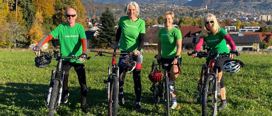 Vier Personen in grünen Shirts und Helmen stehen auf einem Rasenfeld mit ihren Fahrrädern. Dahinter sind Bäume und Berge.