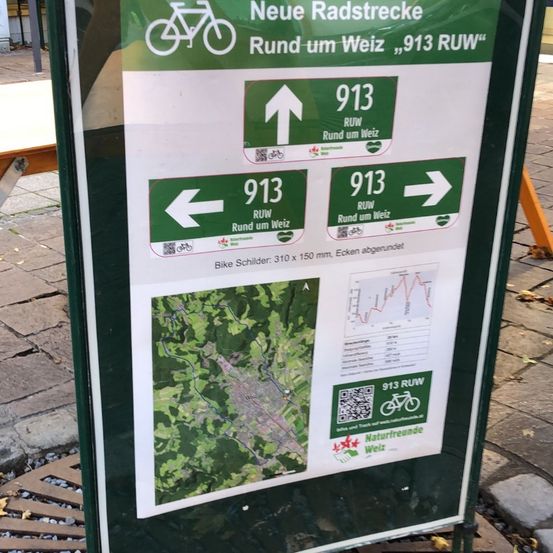 Ein grünes Schild mit einem Fahrradsymbol und Pfeilen in verschiedene Richtungen. Es steht 'Neue Radstrecken Rund um Weiz, 913 RUR' und hat eine Karte und etwas Text darunter. QR-Codes sind vorhanden.