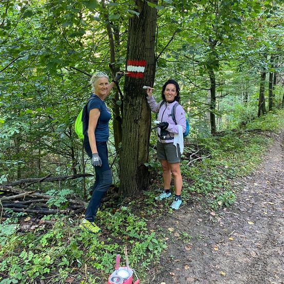 Zwei Frauen im Wald mit Rucksäcken malen einen Baumstamm mit roter und weißer Farbe. Sie lächeln und stehen auf einem Lehmweg.