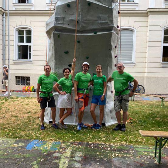 Fünf Personen in grünen T-Shirts stehen vor einer großen Kletterwand und posieren für ein Foto. Dahinter ist ein Gebäude mit mehreren Fenstern zu sehen, und ein Fahrrad ist in der Nähe geparkt.