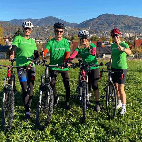 Vier Personen in grünen Radsporttrikots posieren mit ihren Fahrrädern auf einer Wiese, Berge im Hintergrund.