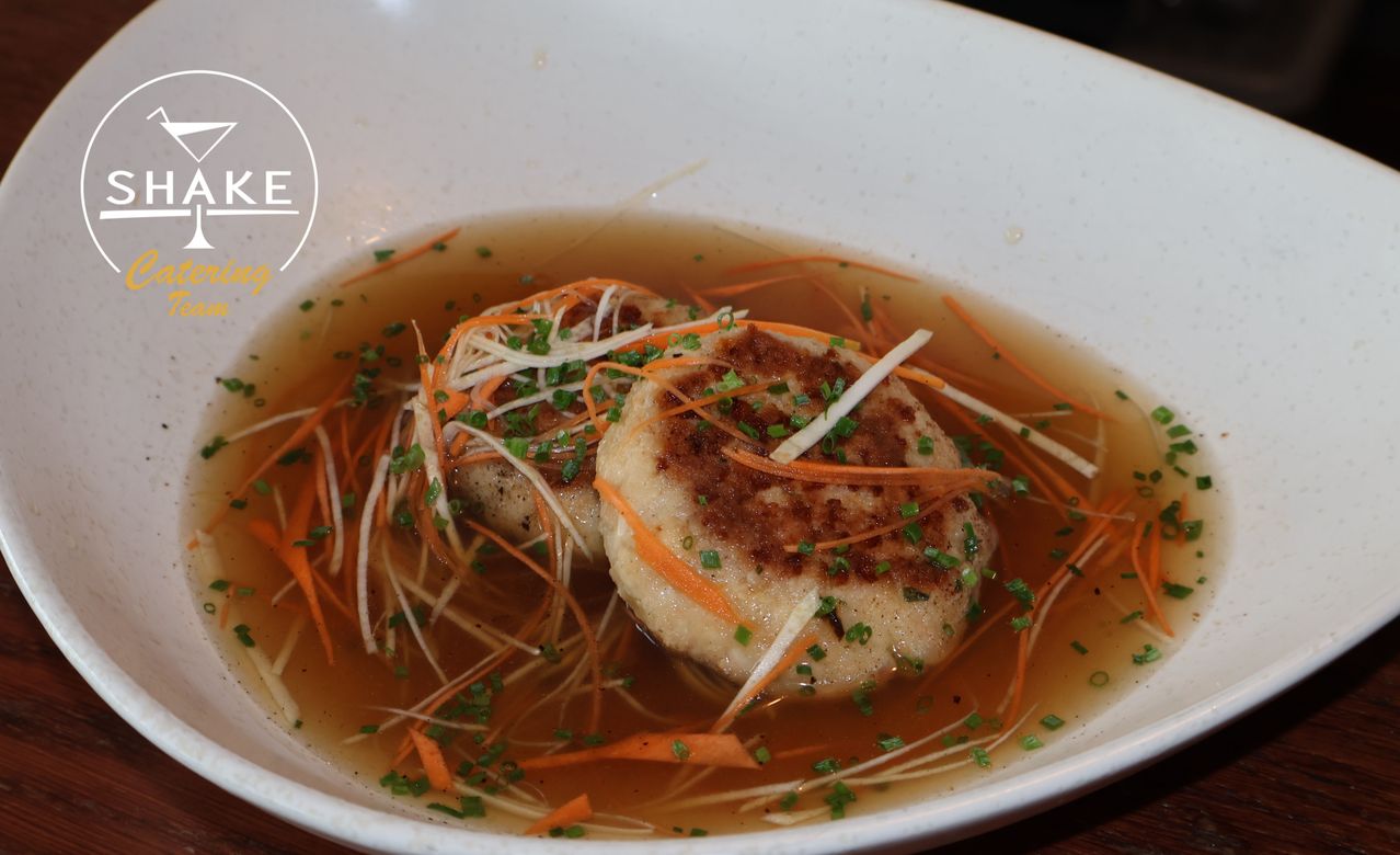 A bowl filled with soup contains two round dumplings topped with green herbs and shredded carrots. The soup is brown and appears to be a broth.