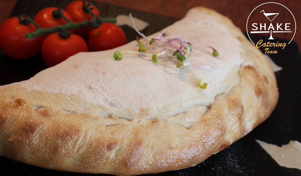 A baked pastry with a golden-brown crust, topped with sprouts, lies on a dark surface near a bunch of red tomatoes.