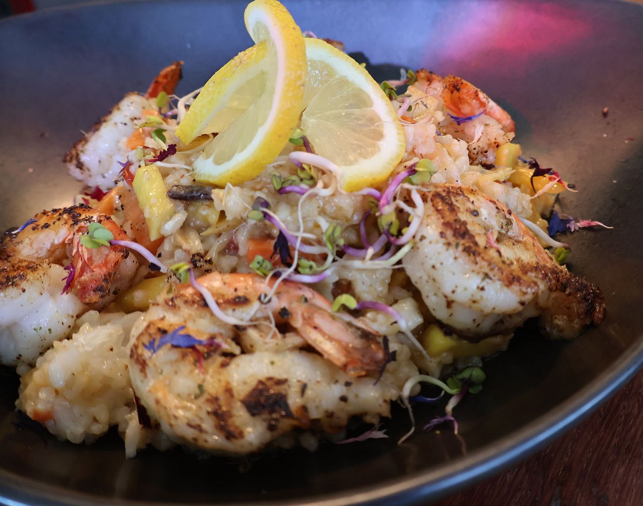 Ein Gericht aus Shrimps-Risotto mit Zitronenscheiben und verschiedenen Garnierungen, serviert auf einem dunklen Teller. Das Gericht ist mit Microgreens und buntem Gemüse garniert.