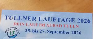 Ein Flyer für das Tullner Lauftage 2026 Event, mit verschiedenen Laufdistanzen und Marathon-Optionen. Es enthält einen QR-Code für die Anmeldung und ist auf einem Holztisch platziert.