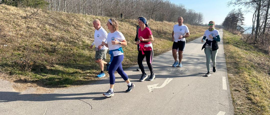Fünf Läufer joggen auf einem Betonweg, umgeben von Grasflächen und Bäumen. Sie tragen weiße Shirts und Sneakers.