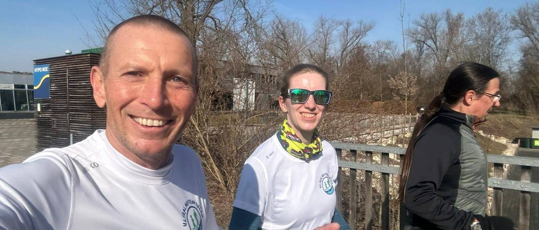 Ein Mann und eine Frau in weißen Shirts lächeln und posieren für ein Foto. Sie stehen neben einem Zaun mit einem Baum im Hintergrund.