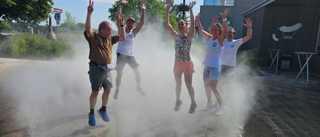 Fünf Personen springen in der Luft und erzeugen eine Nebelwolke. Sie alle lächeln und scheinen Spaß zu haben. Die Umgebung ist im Freien, mit einem Gebäude und Bäumen im Hintergrund.