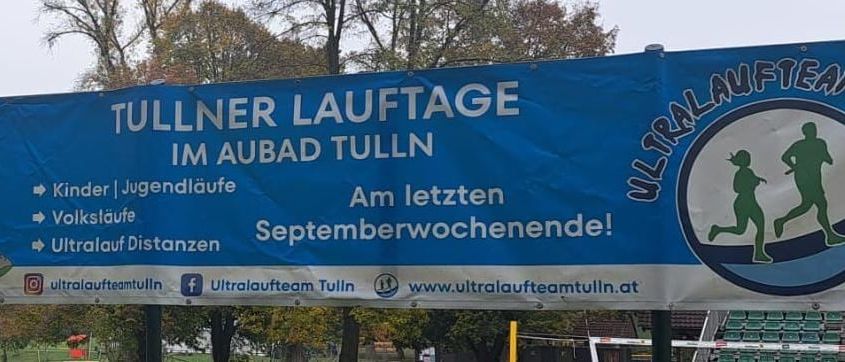 Ein blaues Banner, das für Tullner Lauftage in Aubad Tulln wirbt. Das Banner zeigt Läuferfiguren, Veranstaltungsdetails und Kontaktinformationen. Im Hintergrund sind Bäume und ein Zaun zu sehen.