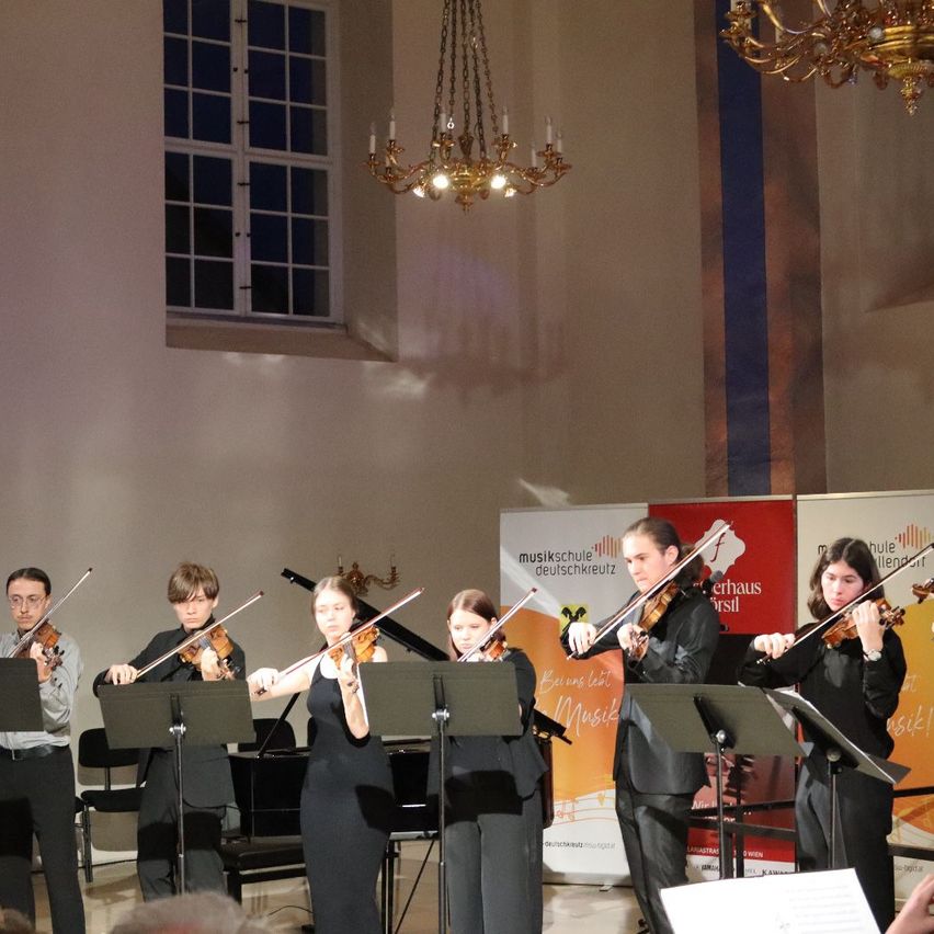 Eine Gruppe von Geigern spielt in einem Raum mit einem Klavier. Sie sind in Schwarz gekleidet und halten Geigen. Eine Kronleuchter hängt von der Decke. Ein Banner ist an der Wand hinter ihnen.