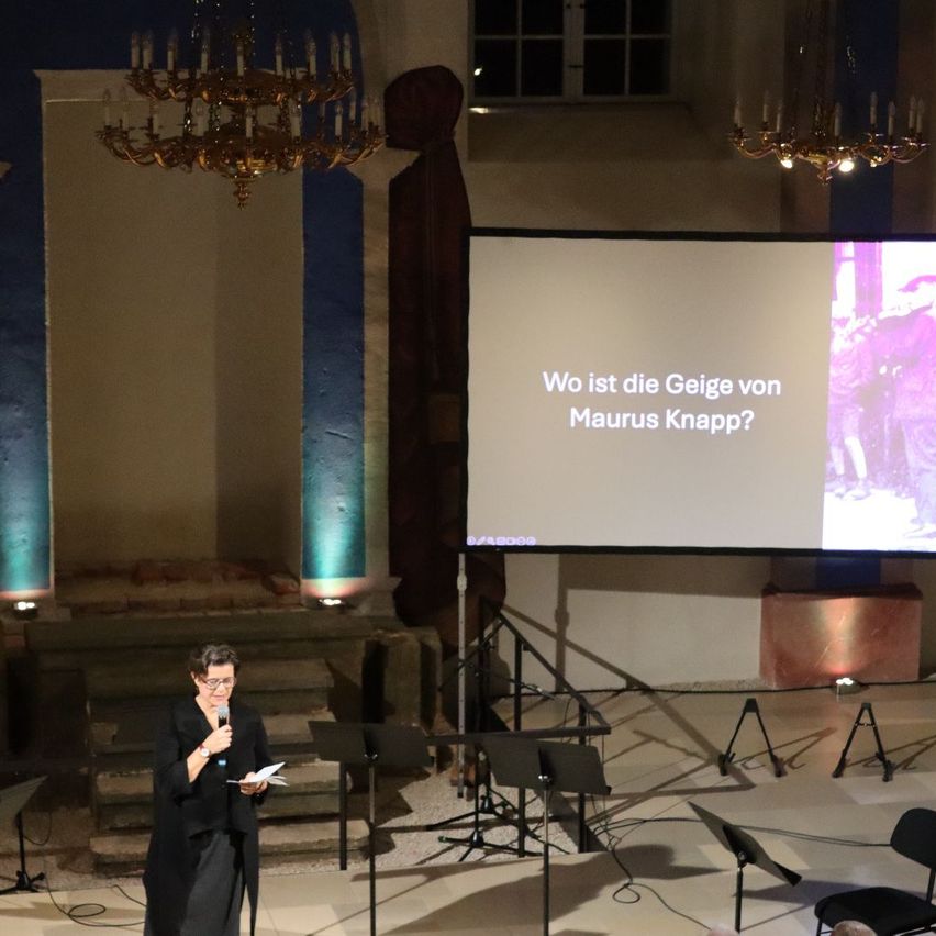 Eine Frau steht auf der Bühne, hält ein Mikrofon und liest von einem Papier. Eine Beamerleinwand zeigt die Frage 'Wo ist die Geiger von Maurus Knapp?' mit einem Bild eines Mannes.