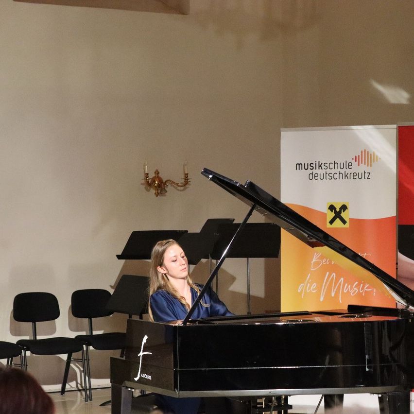 Eine Frau sitzt an einem Flügel mit einem Mikrofonstativ vor ihr. Sie trägt ein blaues Kleid und konzentriert sich auf das Klavier. Hinter ihr steht ein Banner mit der Aufschrift 'Beruf die Musik'. Links sind Stühle in Reihen aufgestellt.