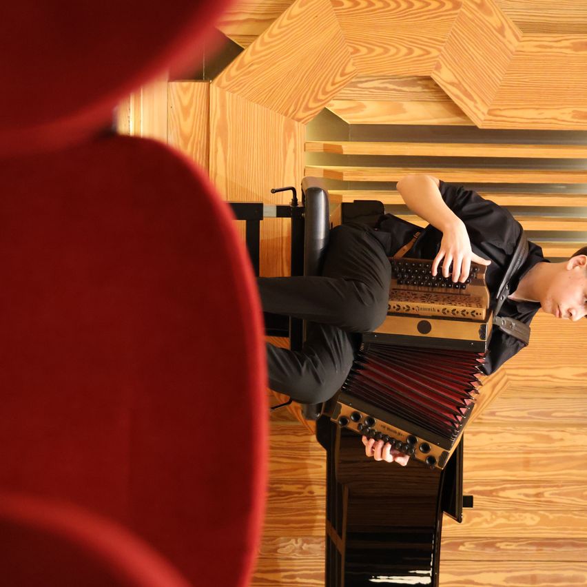 A person playing an accordion with black clothing and a wooden wall in the background.