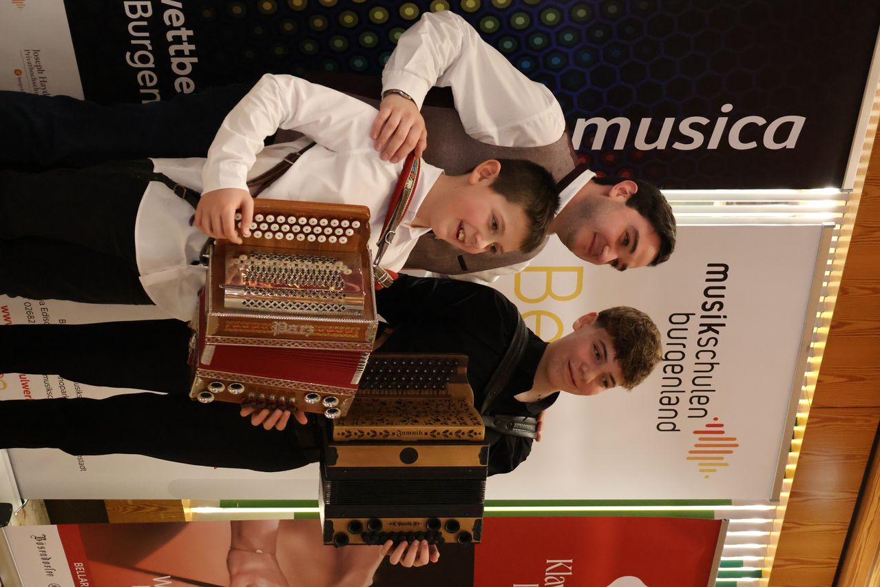 Two young men are posing with an accordion, smiling and holding it in front of them. A banner behind them reads 'Burgenland Musikschulen'.